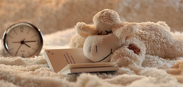 stuffed animal laying on a book in bed as if it's reading next to a clock