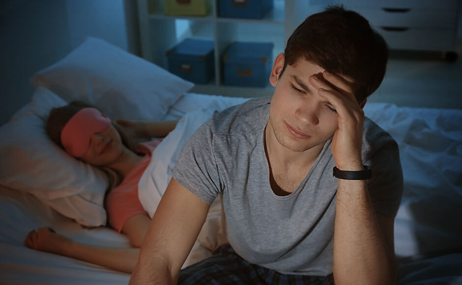 man sitting up in bed in the middle of the night as his girlfriend sleeps peacefully behind him