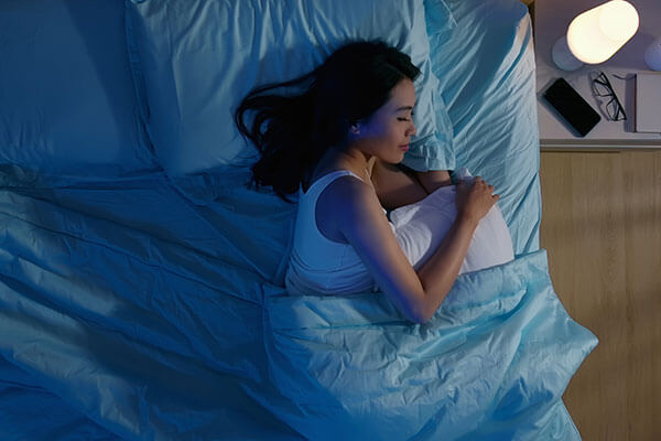 A woman with dark hair peacefully sleeping on her side in a bed with blue sheets and blanket