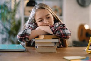 A young girl with her eyes closed, looking sleepy, resting her cheek on her folded arms on a stack of books