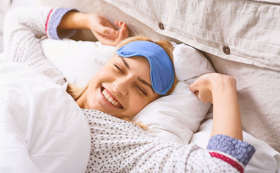 woman waking up and stretching while laying in bed with her eye mask pushed up to her forehead