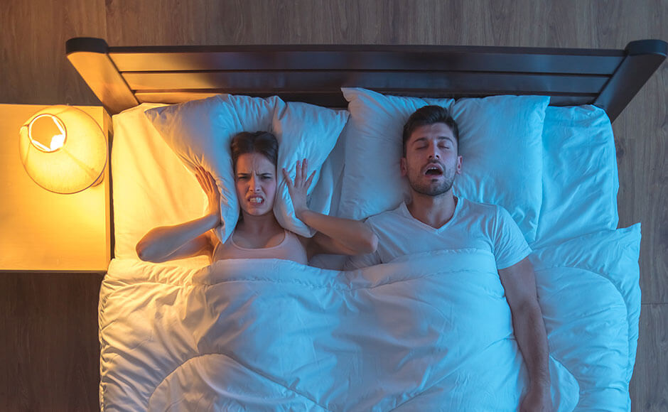 man lays on his back snoring in bed as his wife holds the pillow around her ears to block out the noise